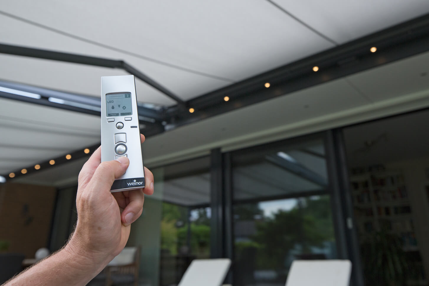 Hand holding a remote control for outdoor shading, with a view of glass sliding walls and a modern terrace.