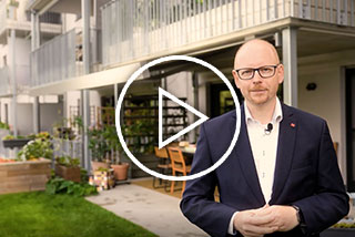 Mann mit Brille steht vor einem modernen Gebäude mit Garten und Terrasse. Spielt auf ein Video an.