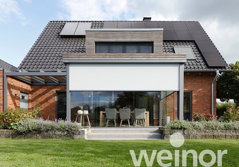 Moderne Fassade eines Hauses mit Markise, Glasfront und grüner Terrassensituation im Vordergrund. 