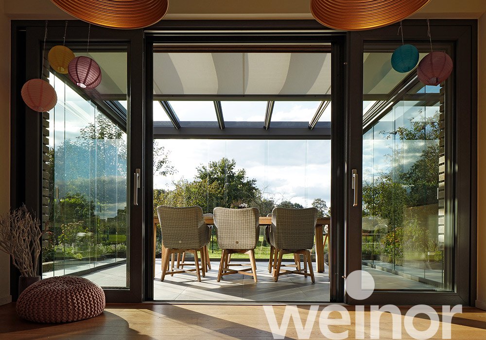 Innenansicht eines Wintergartens mit Glas-Schiebewänden, Tisch und Stühlen, modernem Design und natürlichem Licht. 