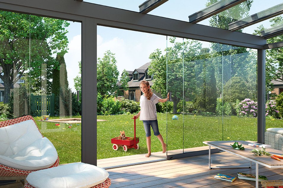 Espace de vie extérieur avec une structure en verre, un jardin verdoyant, et une femme avec un jouet rouge.