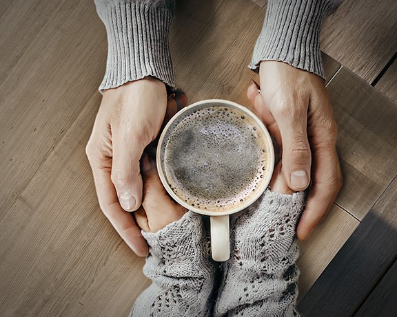Twee handen houden een koffiekopje vast, omringd door een houten tafel. 