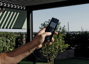 Une main tenant une télécommande avec un fond de verdure et un ciel ensoleillé. 