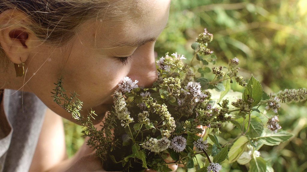 Een vrouw snuffelt naar een bosje met groene en paarse bloemen in een natuurlijke omgeving.