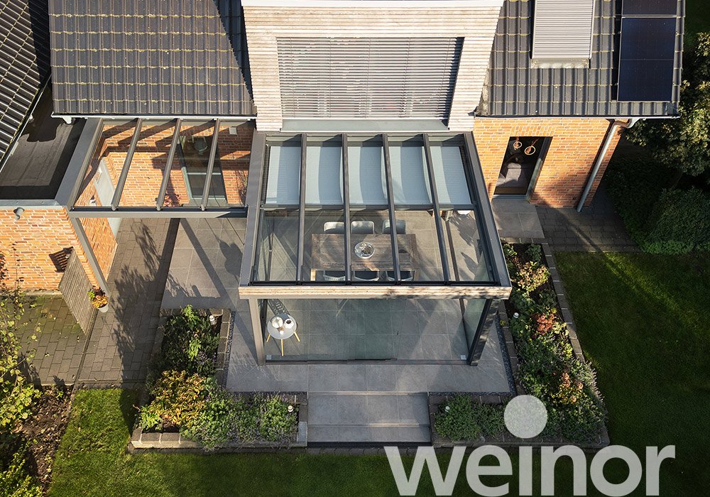 Überdachung mit Glas und Terrassensituation, umgeben von Grünflächen und Pflastersteinen. 