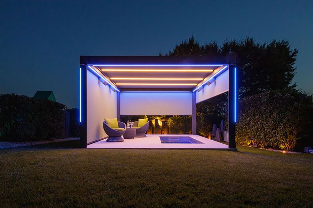 Moderne Überdachung mit LED-Beleuchtung, zwei Sitzmöbeln und Pool in einer grünen Umgebung bei Nacht.
