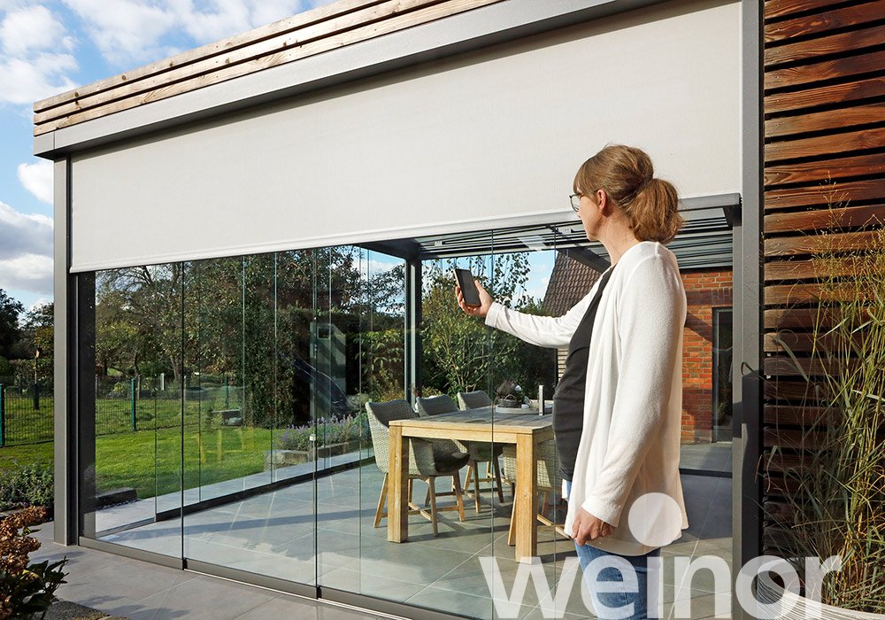Frau steht vor einem Glas-Schiebewand mit einem Smartphone, Blick auf eine Terrassensituation. Dominante Farben: Grau und Blau. 