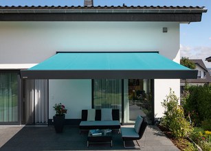 Terrassensituatie met een turquoise zonnescherm boven een zithoek aan een gebouwse gevel. 