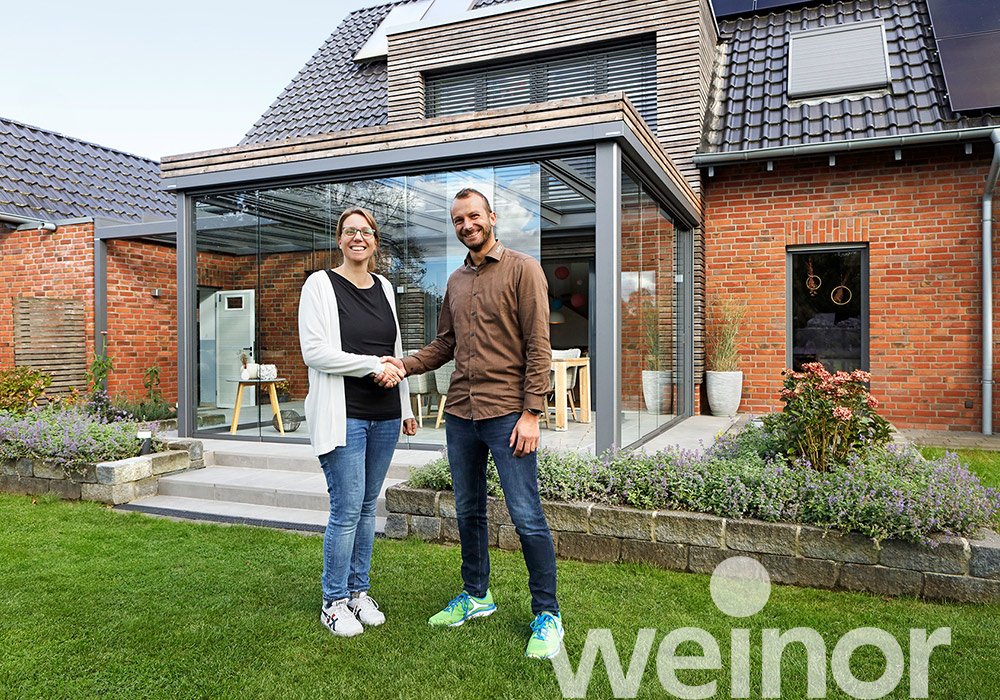 Zwei Personen schütteln sich die Hände vor einem modernen Haus mit Glasüberdachung und gepflegtem Garten. 