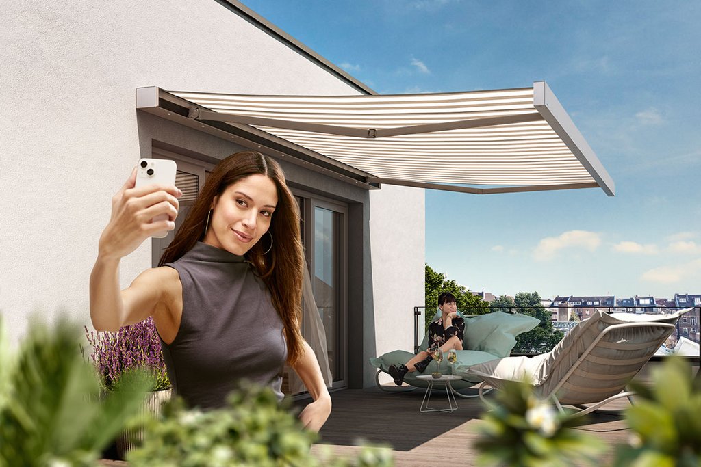 Eine Person macht ein Selfie auf einer Terrasse, im Hintergrund eine Markise und Pflanzen. Himmel mit Wolken.