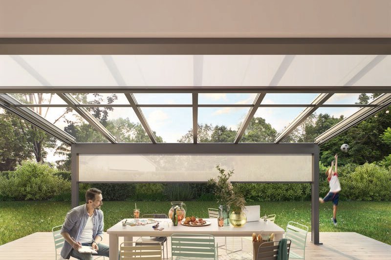 Tafel met stoelen onder een overkapping, groene tuin op de achtergrond en een kind dat speelt.