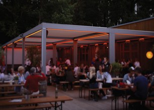 Een moderne overkapping met warme verlichting, vol mensen en houten tafels in een levendige omgeving. 