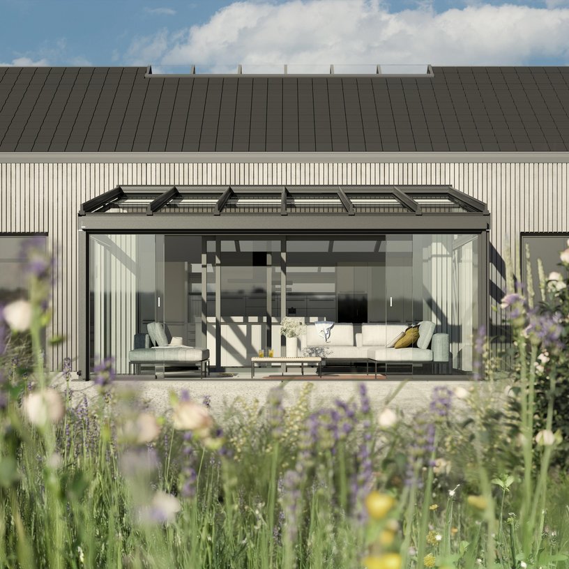 A modern facade with a glass extension and seating area, surrounded by lush greenery.