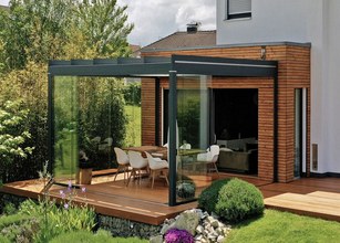 Moderne overkapping met glaswanden en houten terras bij een huis, omringd door groen. 
