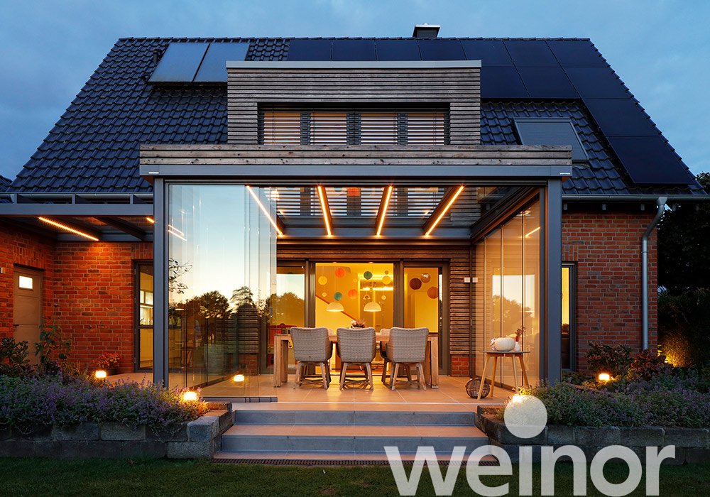 Moderne Hausfassade mit Glas-Schiebewänden, Terrassensituation und warmem Licht bei Abenddämmerung. 