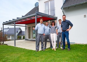 Groep mensen poseert onder een overkapping met een rood doek in een groene tuin. 