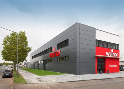 Modernes Gebäude in Grau mit roten Akzenten und großen Fenstern, umgeben von Bäumen und einer gepflasterten Fläche.