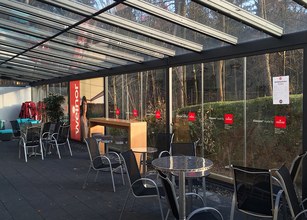 terras met glazen overkapping, stoelen en tafels. Achtergrond met bomen en groene struiken. 