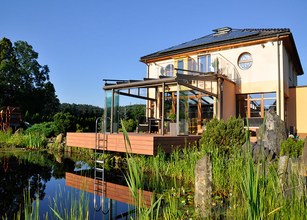 Moderne woning met een terras en een glazen wintergarten, omringd door groen en waterreflectie. 