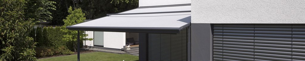 Markise in grauem Stoff, montiert an einer modernen Hausfassade mit glatten Oberflächen und grüner Umgebung.