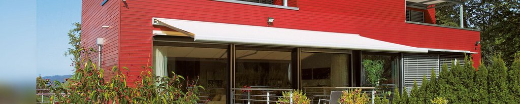 Moderne Architektur mit roter Fassade und Überdachung, umgeben von grünen Pflanzen und Terrassensituation.