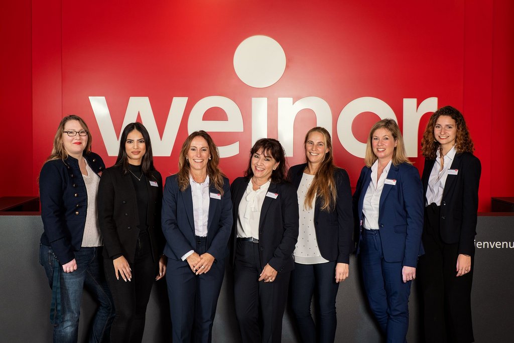 Gruppe von sieben Personen vor einer roten Wand mit dem Schriftzug "weinor". Alle in formeller Kleidung.