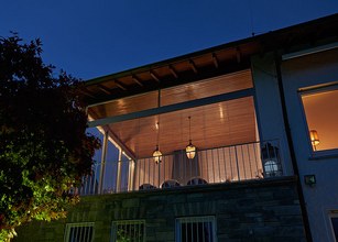 Een terras met overkapping en hangende verlichting bij nacht. Groen van planten aan de zijkant. 
